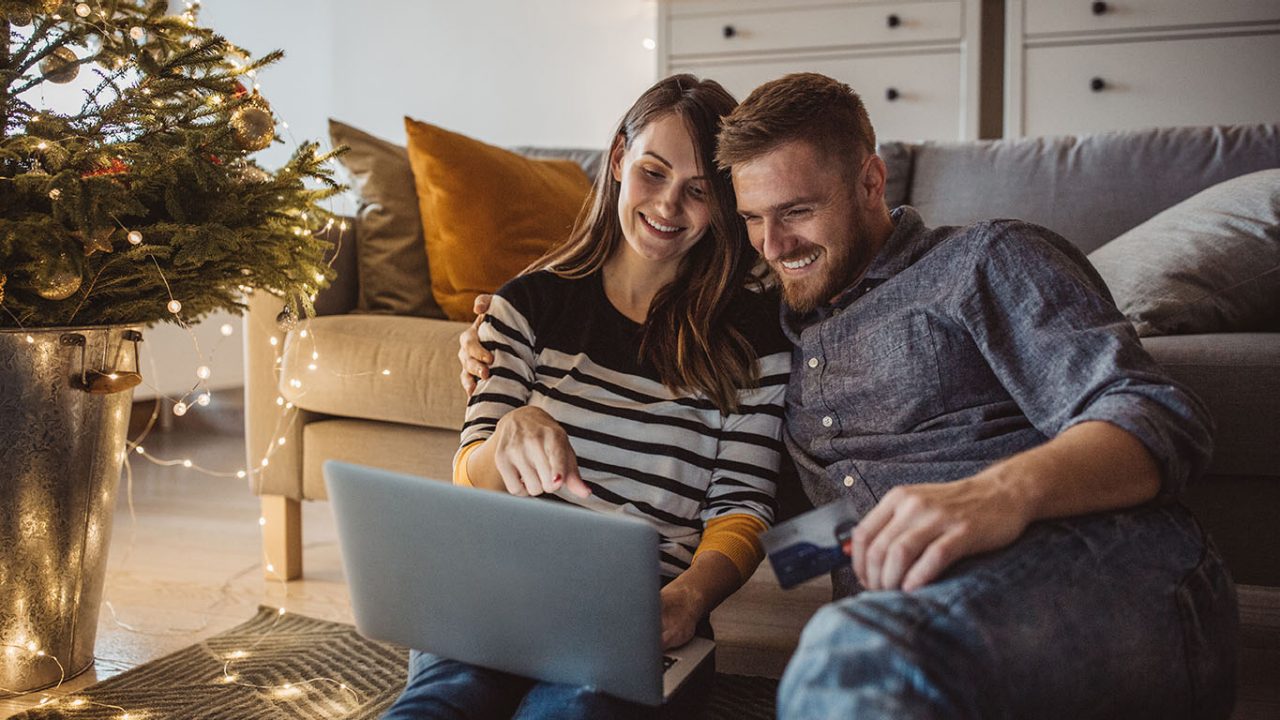 Young couple celebrating Christmas at home. They sitting on floor and shopping online. Home is decorated with Christmas ornaments and lights.

