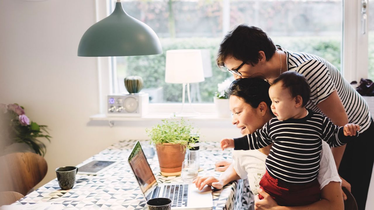 A same-sex couple with their child looking at a laptop in their kitchen.