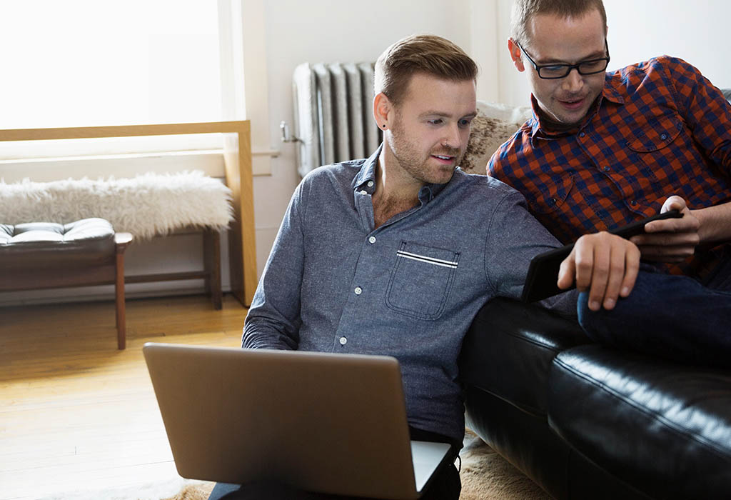 Two young men in a living room, one on a couch, and one on the floor looking at a phone with a laptop.