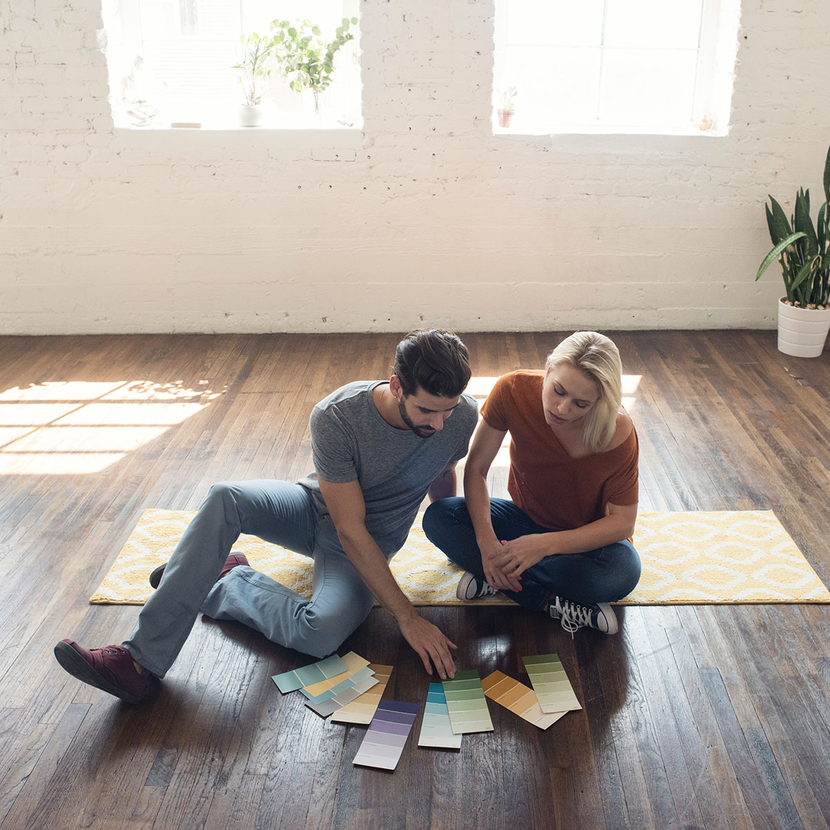 A couple sits together on their hardwood floors in an empty home going through colour samples.