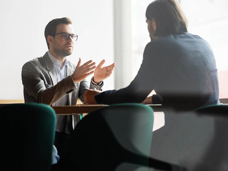 Mid shot image, shot through glass into a meeting room. A man with glasses and wearing a suit is gesturing and talking to a man across the table from him.