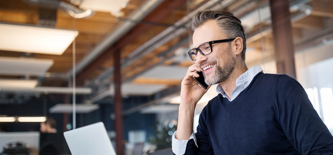 Mid adult business professional sitting at table with laptop and talking on mobile phone. Businessman working in office.