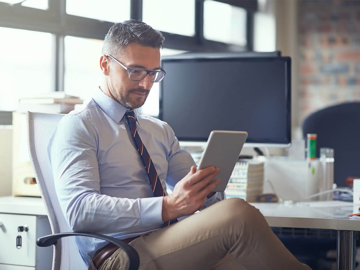 A middle aged man with glasses and tie looking at a tablet while sitting at a modern office desk.