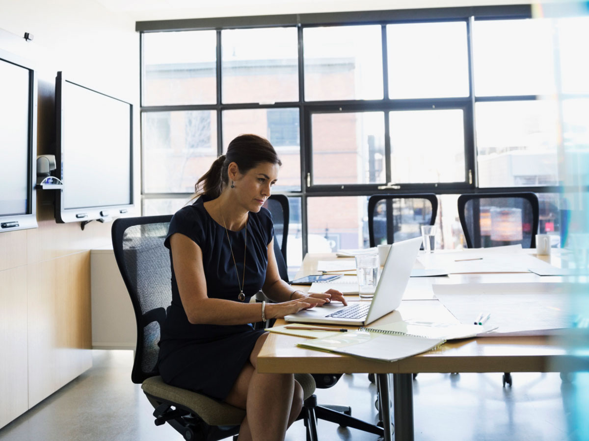 A professionally dressed woman typing on a laptop in a brightly lit boardroom.