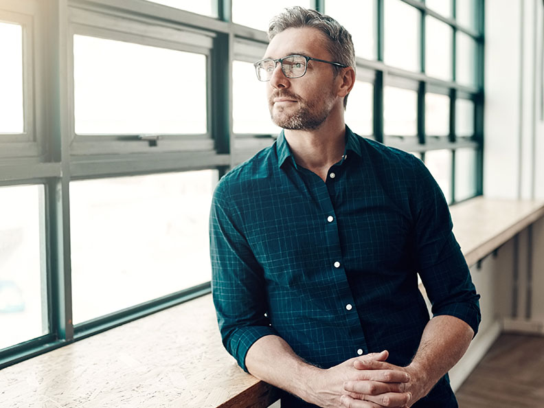 A middle aged man with glasses in smart casual clothing looking out a window of a modern office