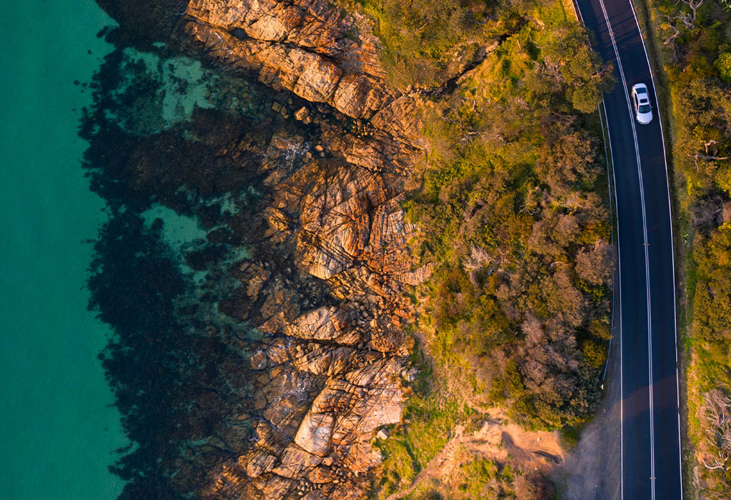 A car driving on the left side of a coastal road, birds eye view, day time.