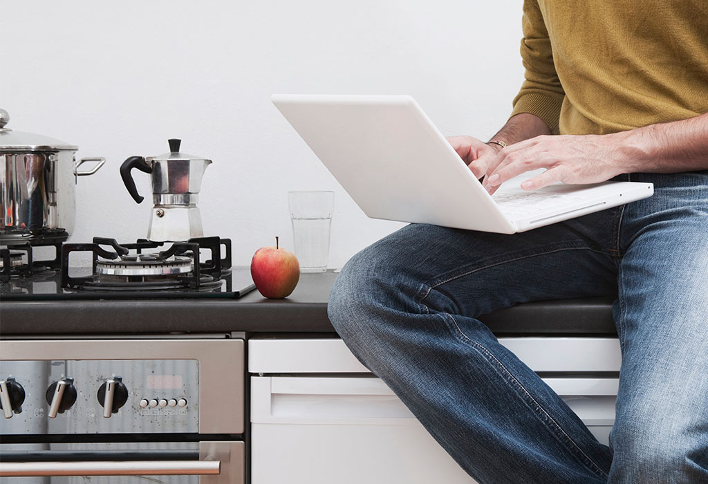 A lower half view of a person sitting on a kitchen counter with their laptop open.