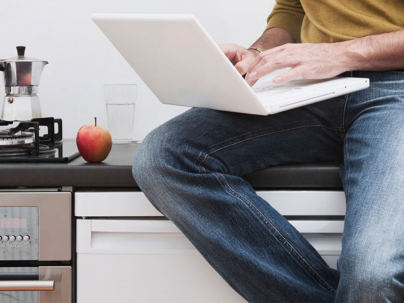 A lower half view of a person sitting on a kitchen counter with their laptop open.