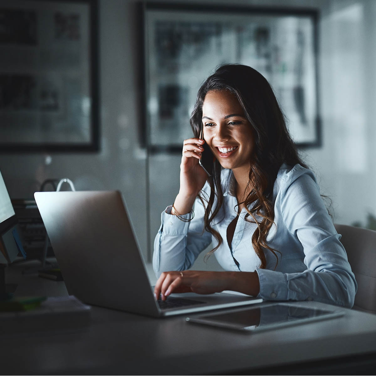 A long haired business woman sitting at a desk speaking to someone on a mobile phone in an office.