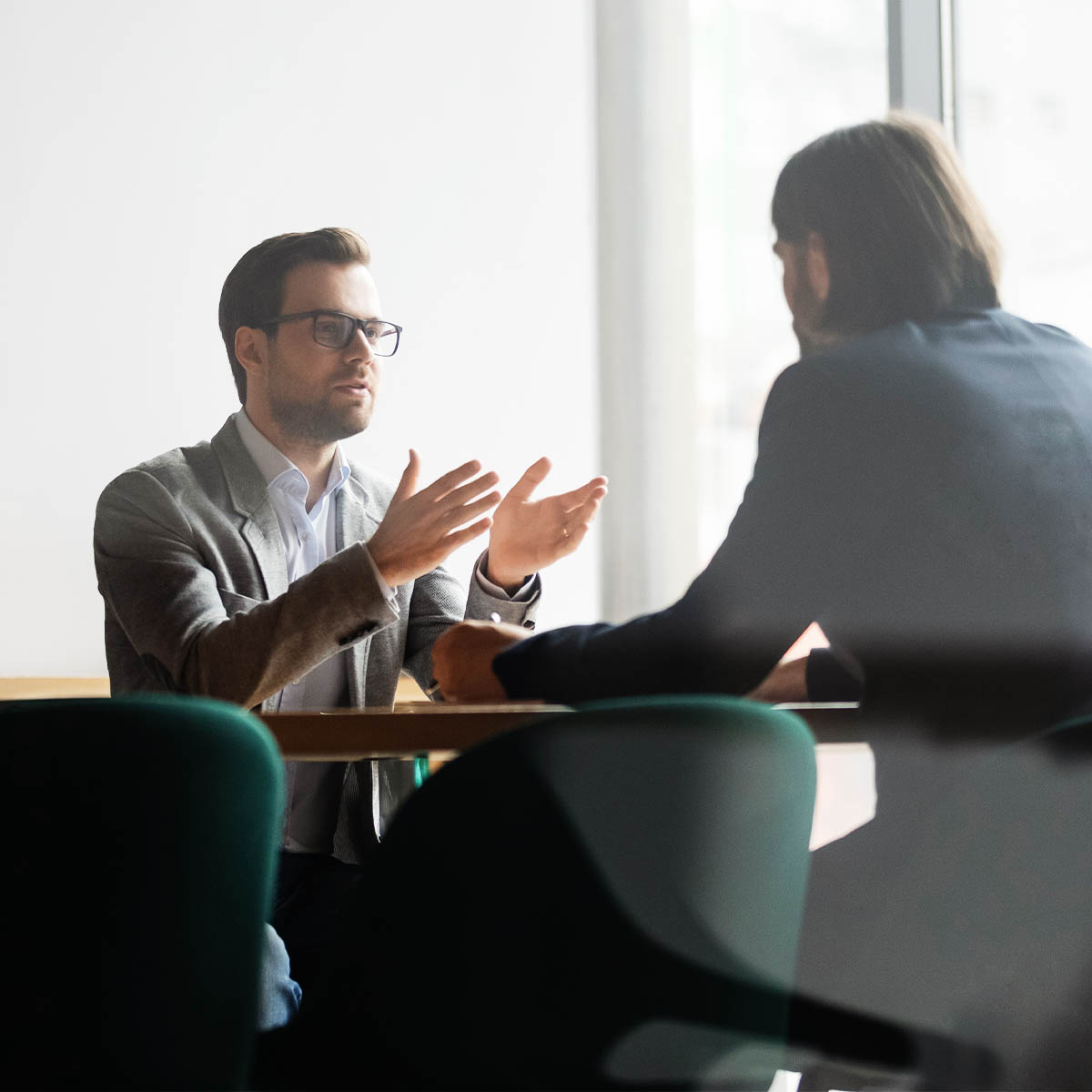 Mid shot image, shot through glass into a meeting room. A man with glasses and wearing a suit is gesturing and talking to a man across the table from him.