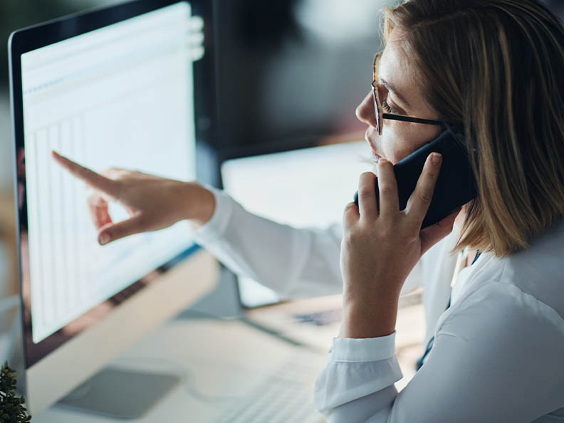 A business woman with glasses in an office, sitting at her desk and on her mobile, points at a computer screen
