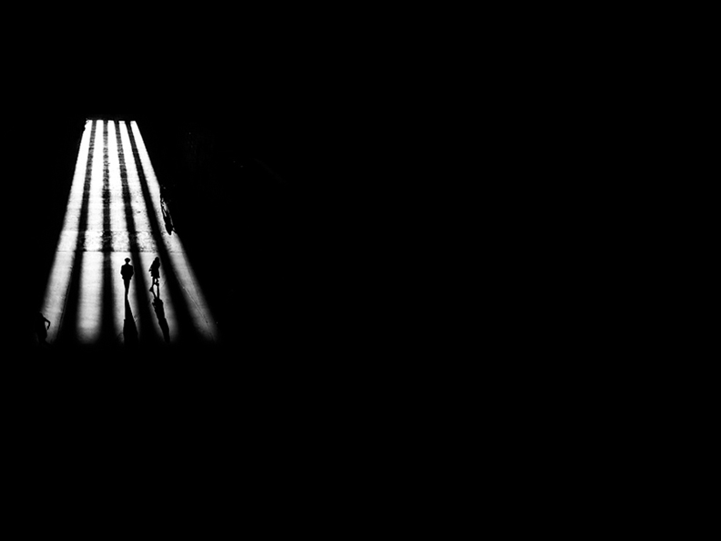 Black and white image. A high angle shot of people walking between light and shadows cast by large pillars.